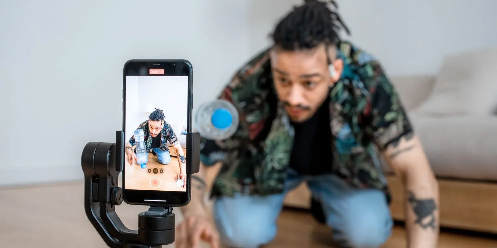 Homem ajoelhado no chão de casa gravando um vídeo de si mesmo com o celular preso num estabilizador