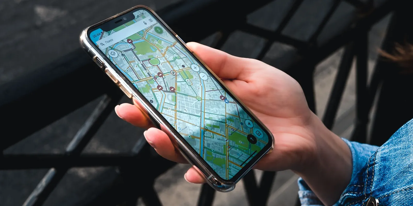 Mão segurando um celular com um aplicativo de mapa aberto na tela, sobre uma grade de metal