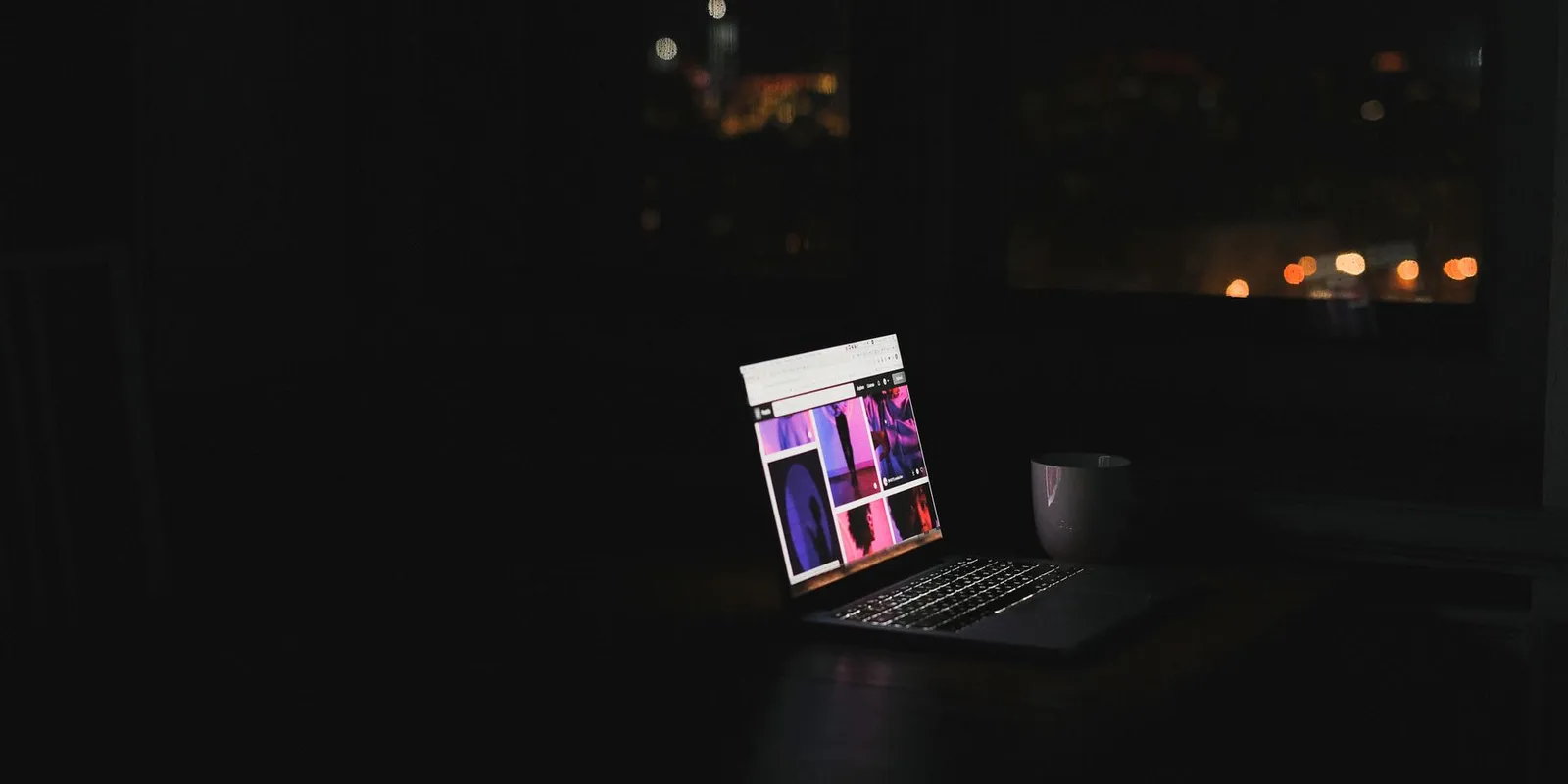 Notebook aceso sozinho numa mesa de um cômodo escuro, com luzes da cidade pela janela