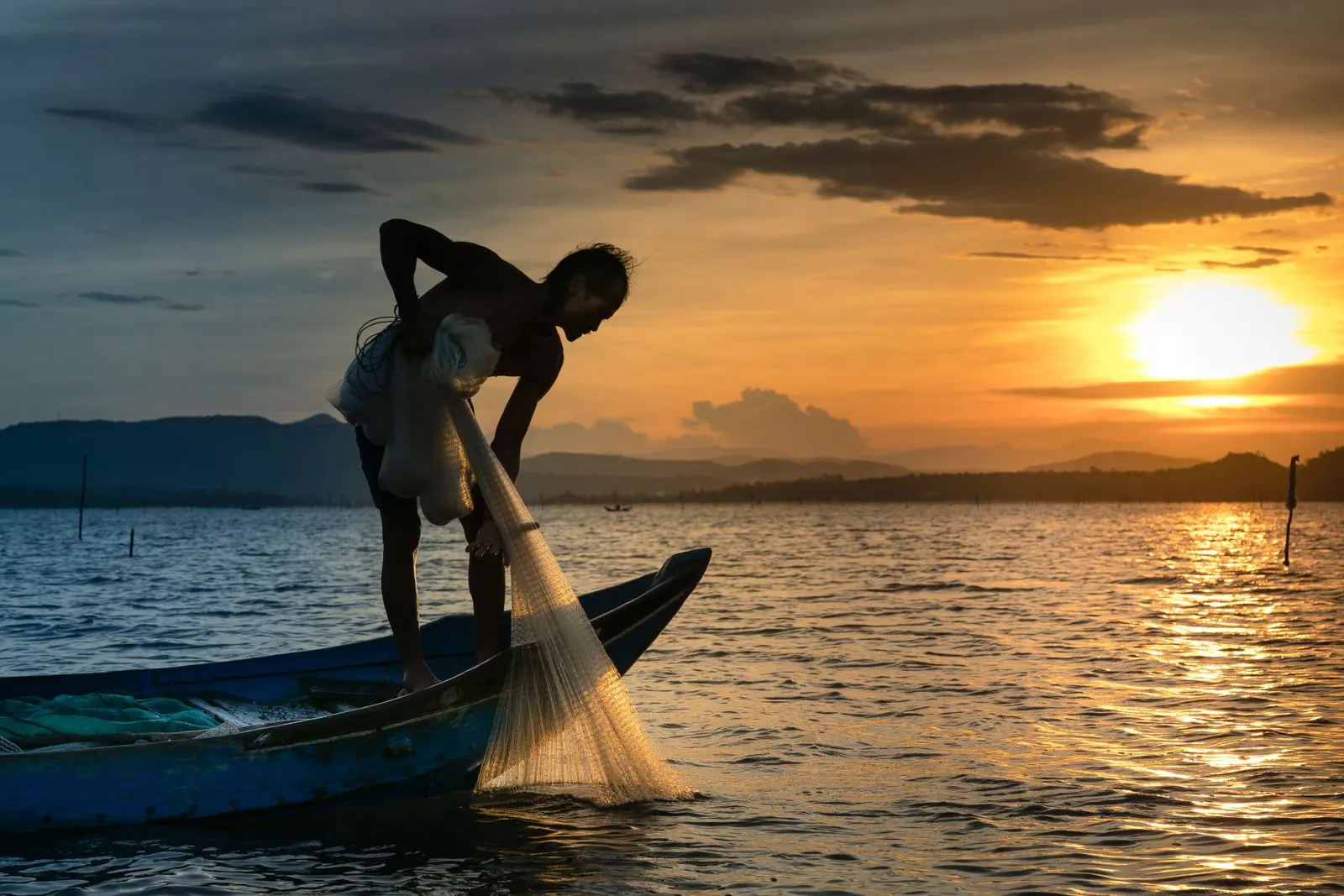 Pescador em pé num barco lançando a rede no mar ao pôr do sol