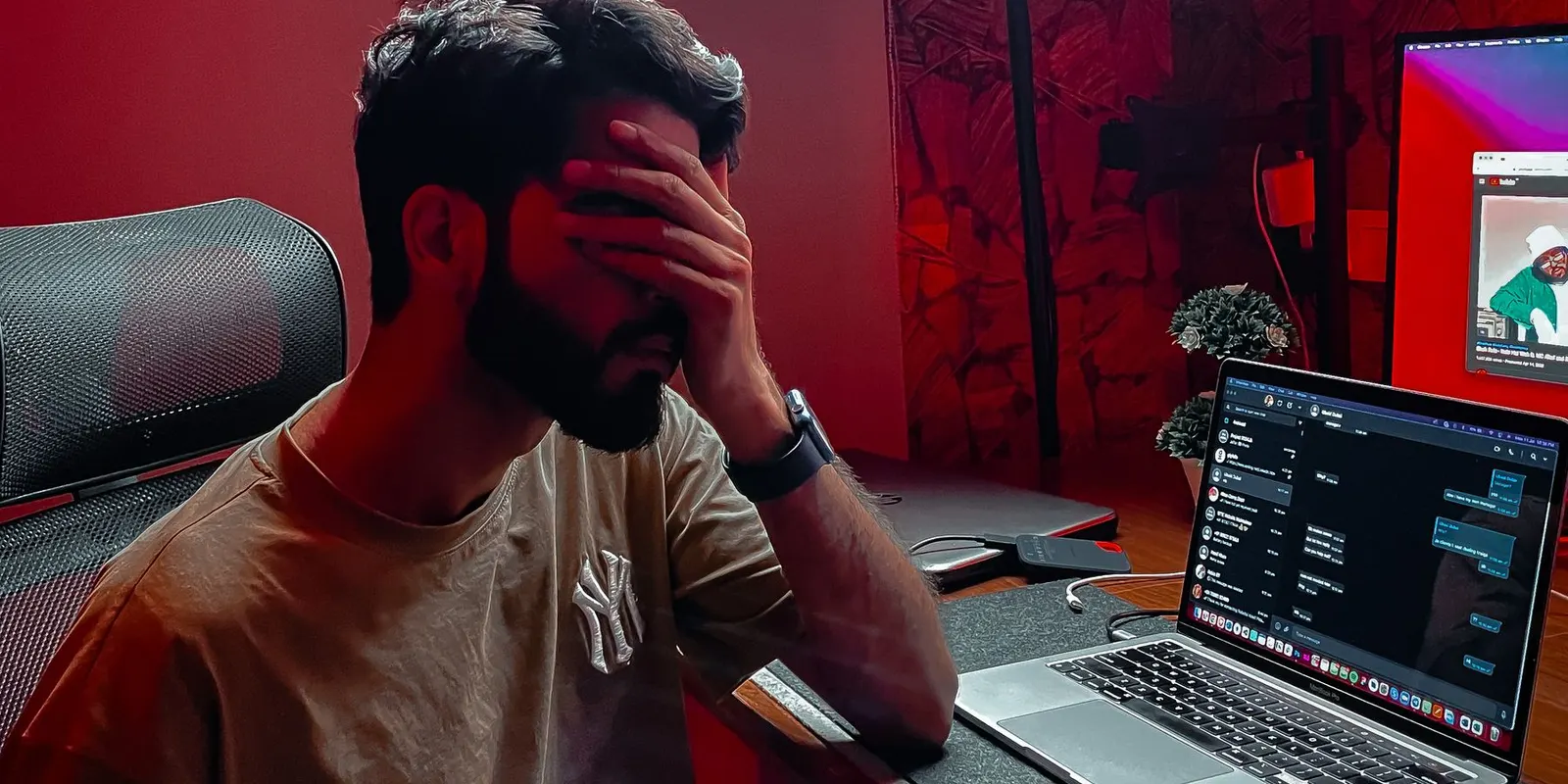 Homem com a mão no rosto, frustrado, sentado na frente de um notebook aberto